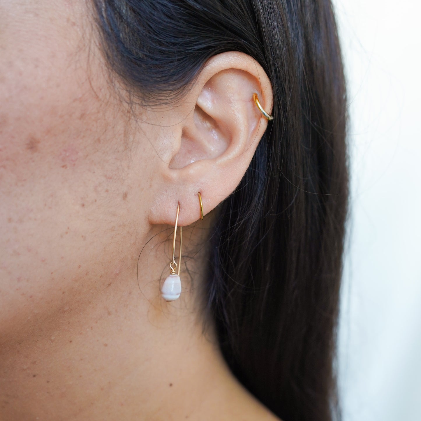 Close-up of an ear wearing gold threader hoop earrings with tiny cone shell on a blurred background