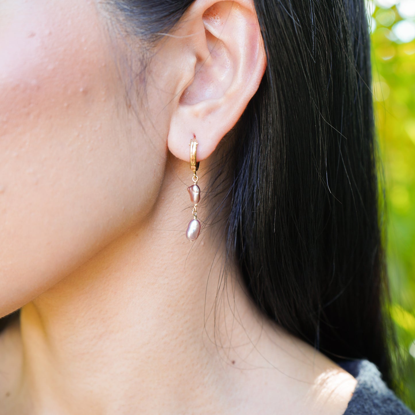 Close-up of an ear wearing a gold hoop earring with dangling pink pearls, blurred green background
