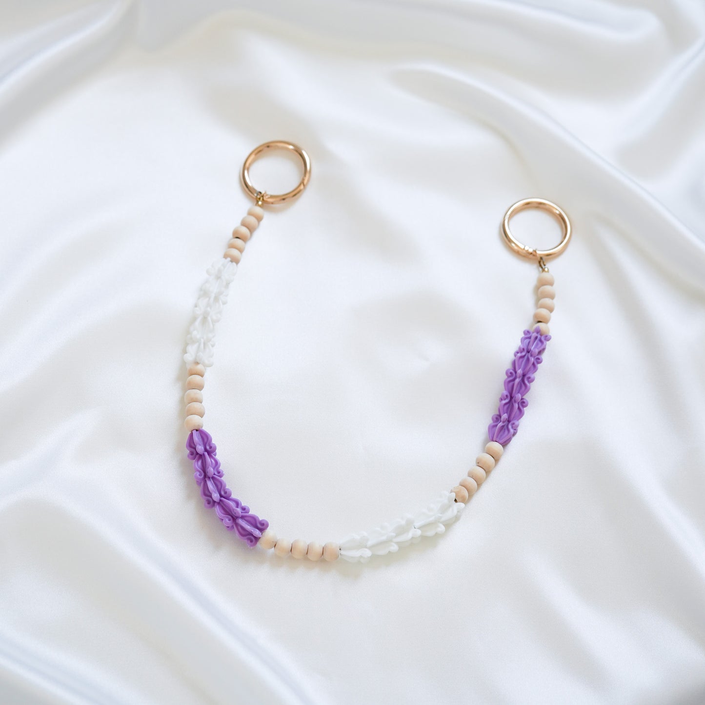 Delicate bag lei featuring purple and white crown flower beads with gold rings on either end on a white fabric background