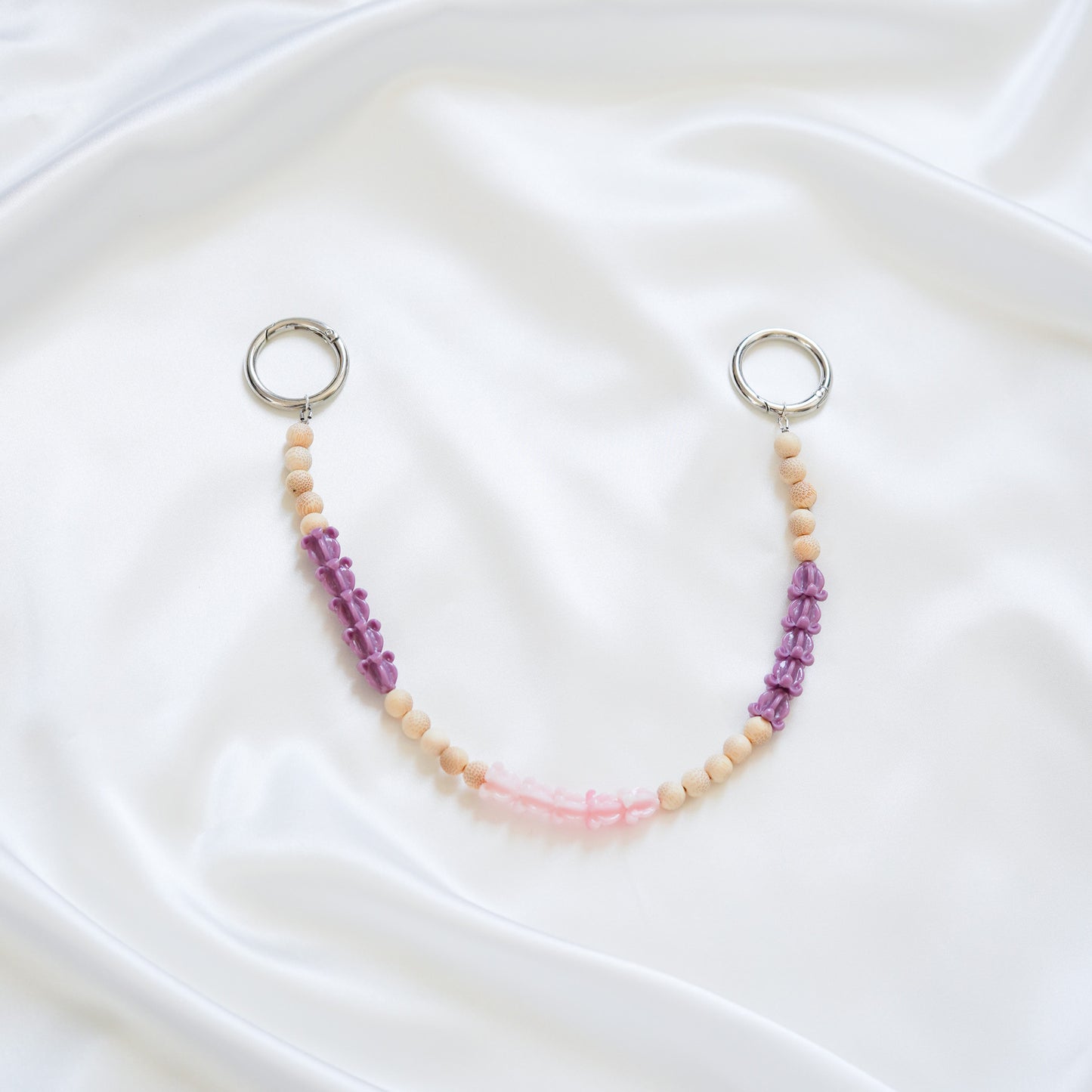 Delicate bag lei featuring purple crown flower, pink crown flower and tan beads in sets of five with gold rings on either end on a white fabric background