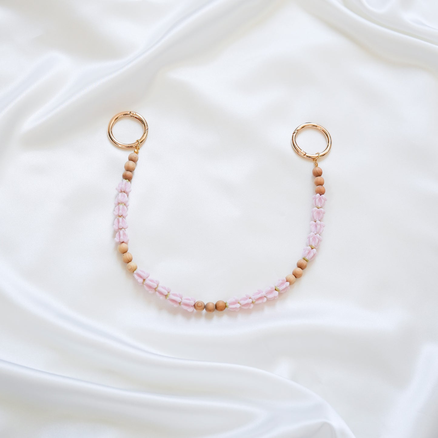 Delicate bag lei featuring pink crown flower and tan beads with gold rings on either end on a white fabric background