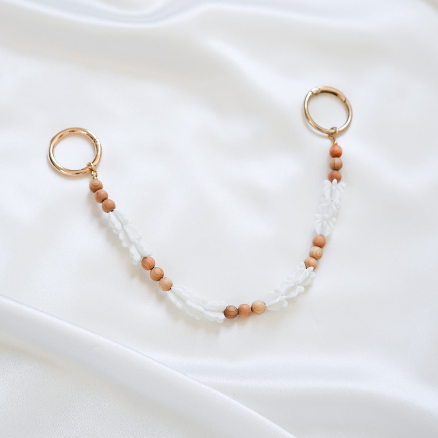 Delicate bag lei featuring white crown flower beads and tan beads alternating in sets of three with gold rings on each end on a white fabric background
