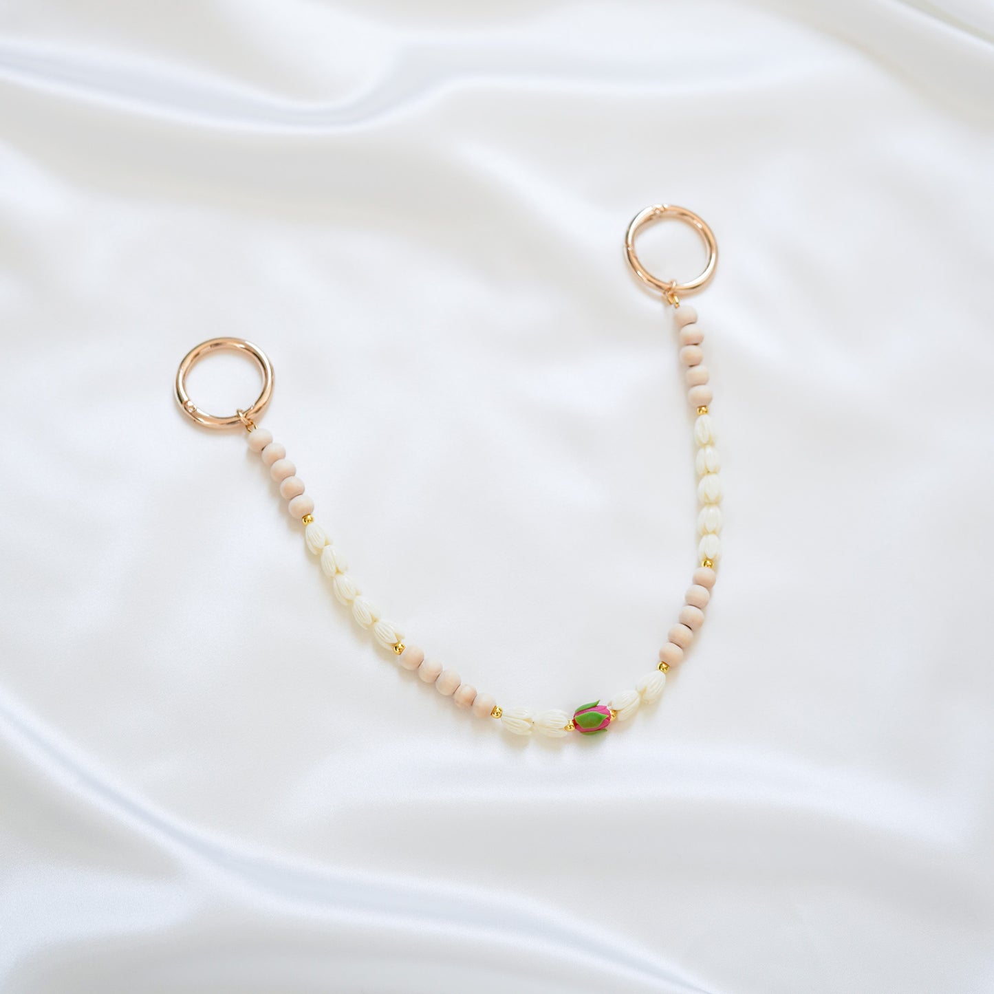 Delicate bag lei featuring white pikake flower beads and light tan beads with a pink rose bead in the middle with gold rings on each end on a white fabric background