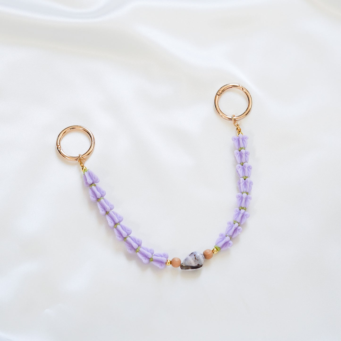 Delicate bag lei featuring purple crown flower and a patterned shell in the middle with tan beads around shell with gold rings on each end on a white fabric background