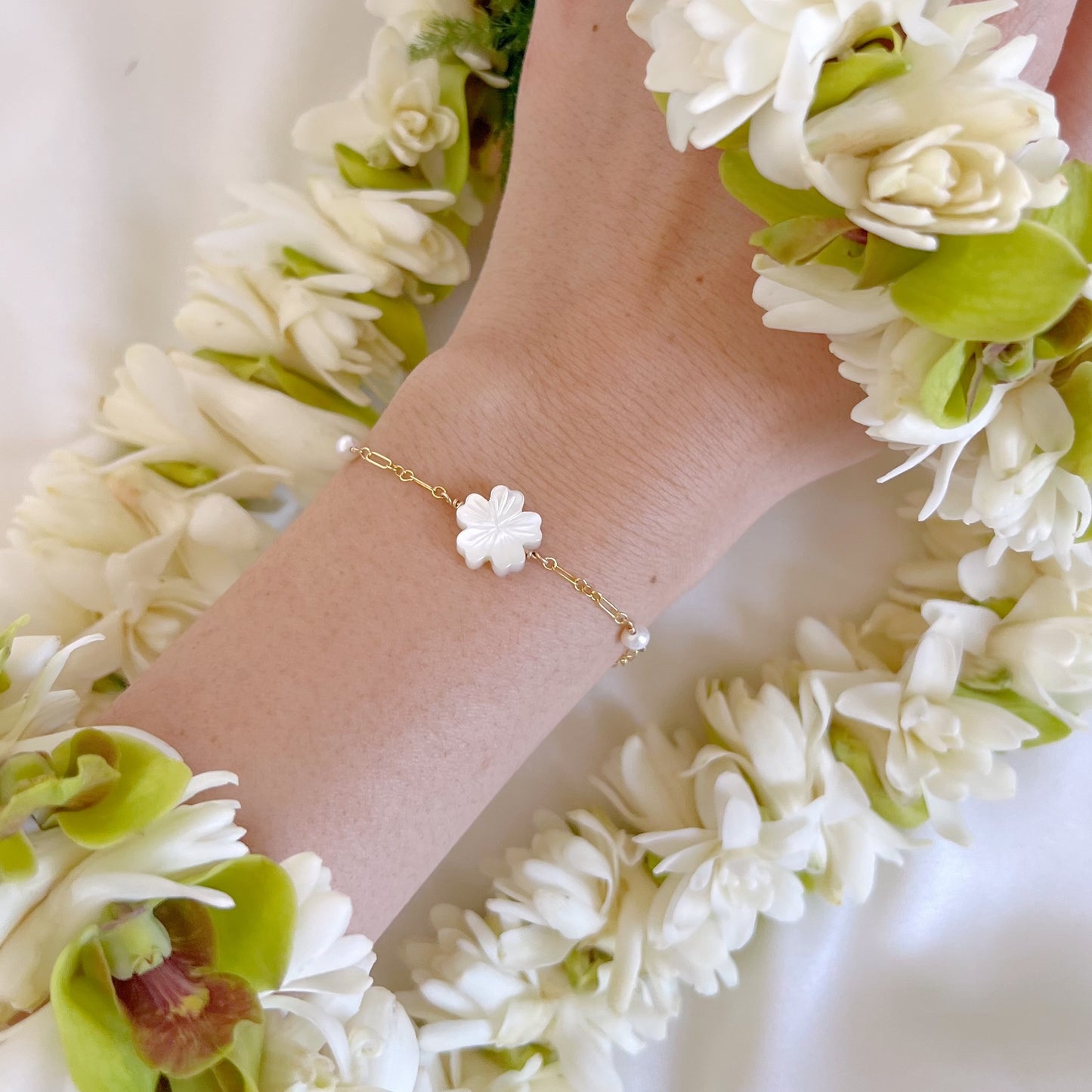 Hand wearing a gold bracelet with a white flower charm and two pearl beads, surrounded by a white and green lei.