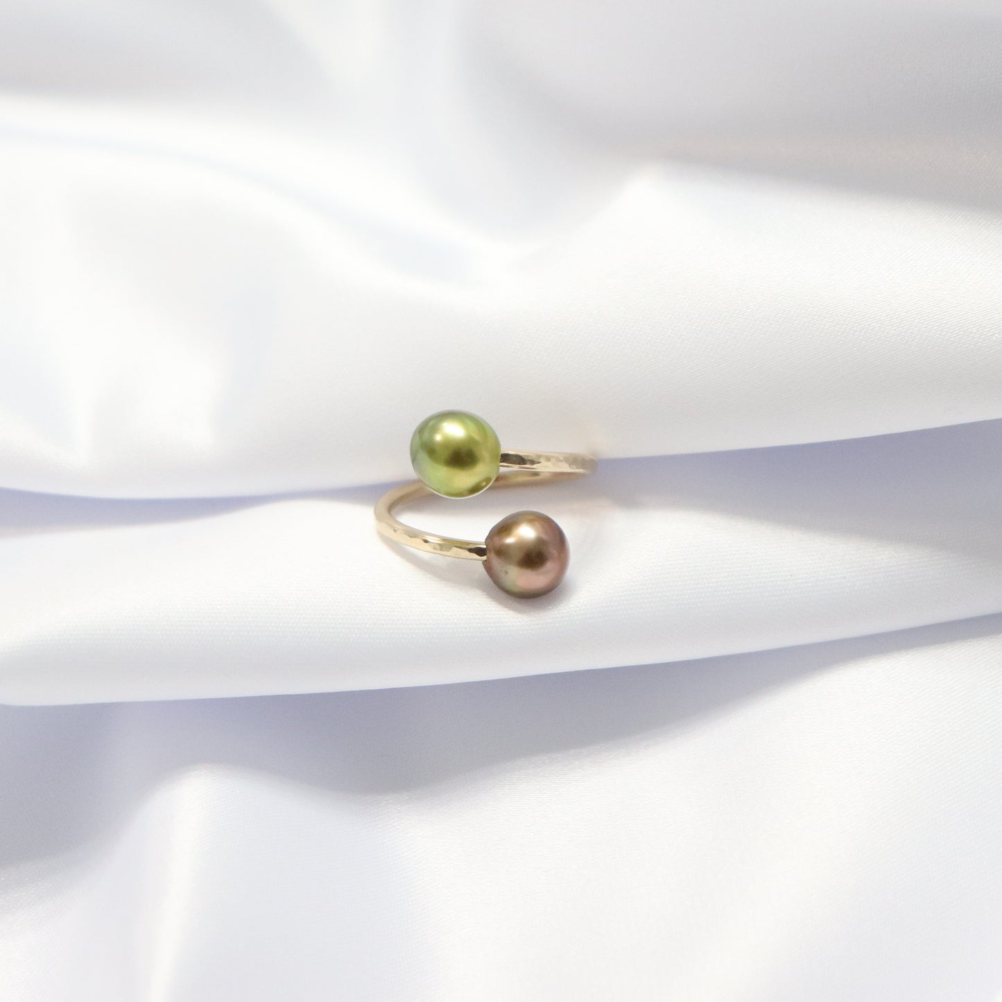 Gold ring with one green and one brown pearl on a white fabric background