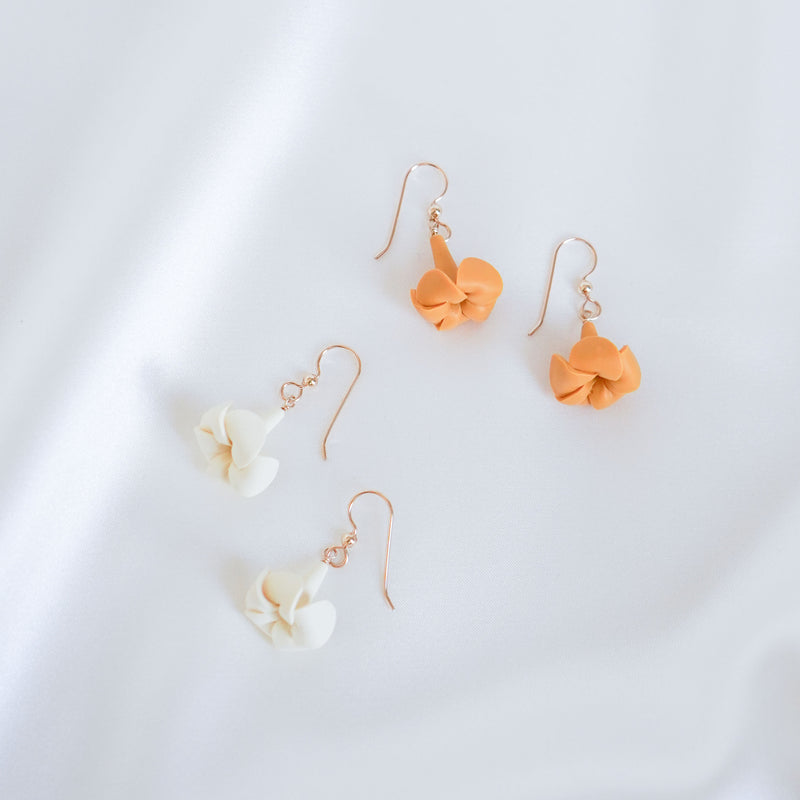 Two pairs of gold  earrings with an orange or white puakenikeni flower pendant on a white background
