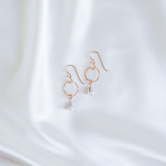 Gold circle drop earrings with tiny cone shell on a white fabric background