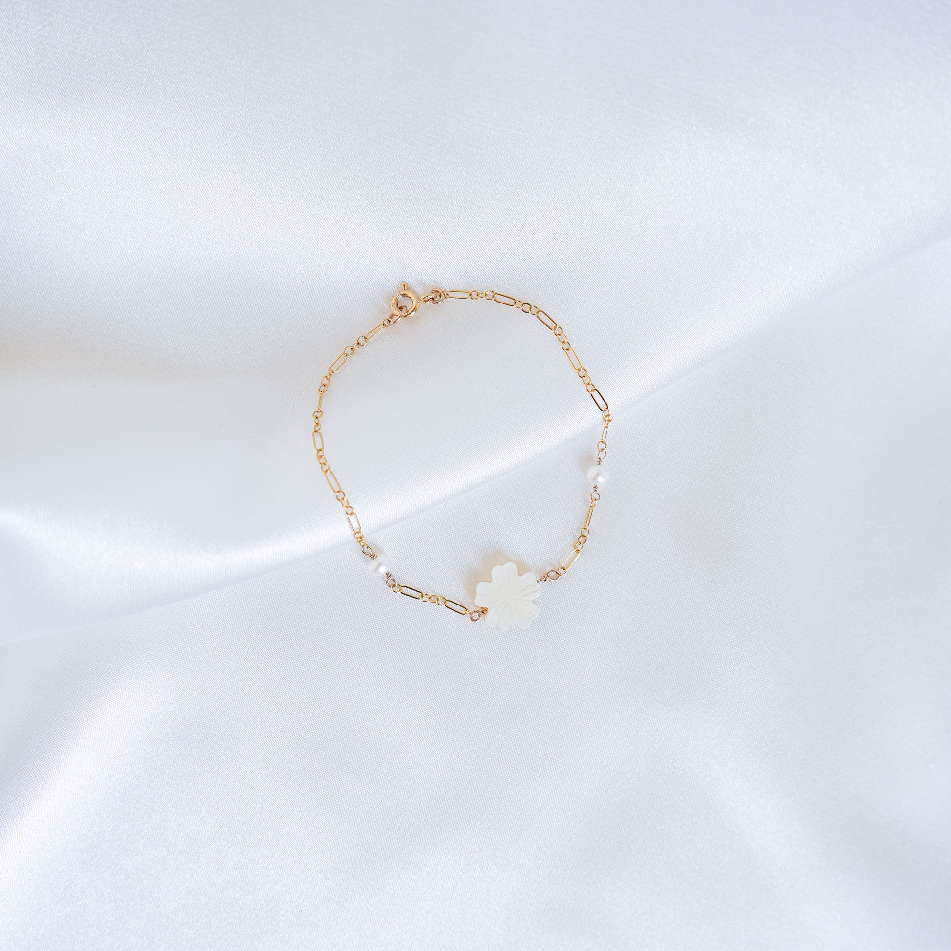 Gold bracelet with a white mother of pearl floral charm and two pearl beads on a white satin background