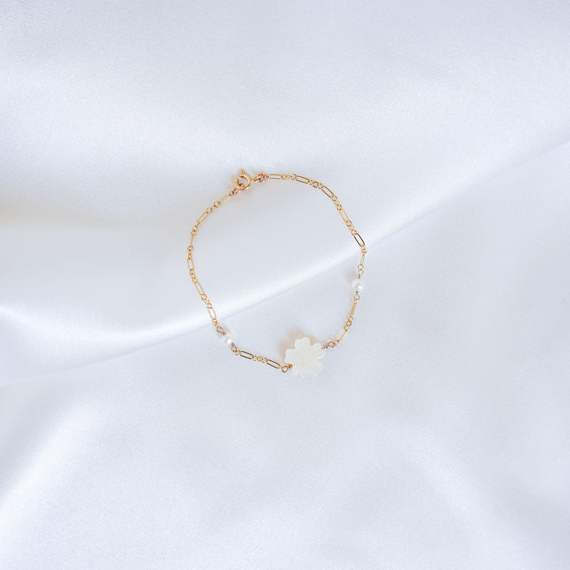 Gold bracelet with a white mother of pearl floral charm and two pearl beads on a white satin background