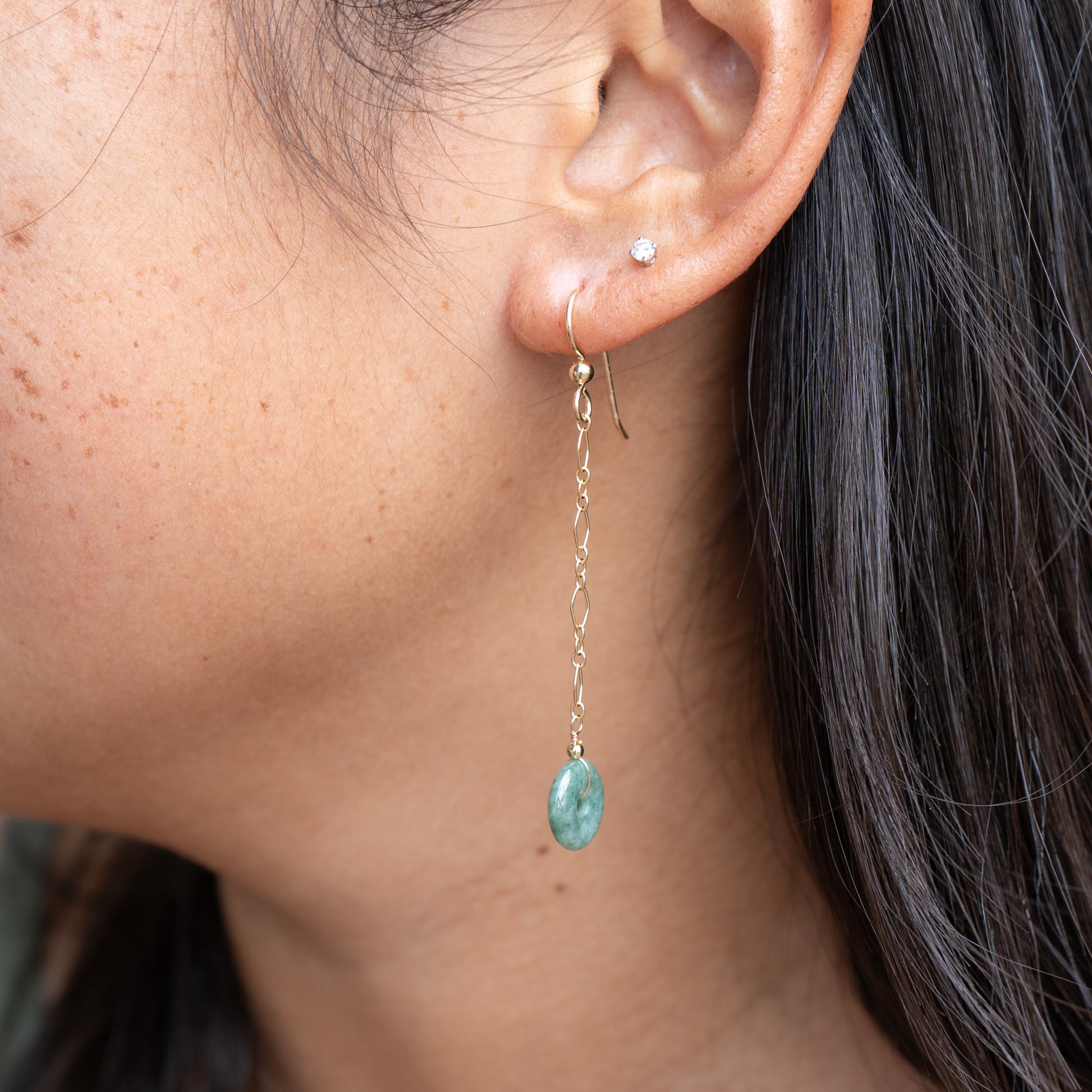Close-up of an ear wearing a gold dangle chain earring with circle green jade with a blurred background
