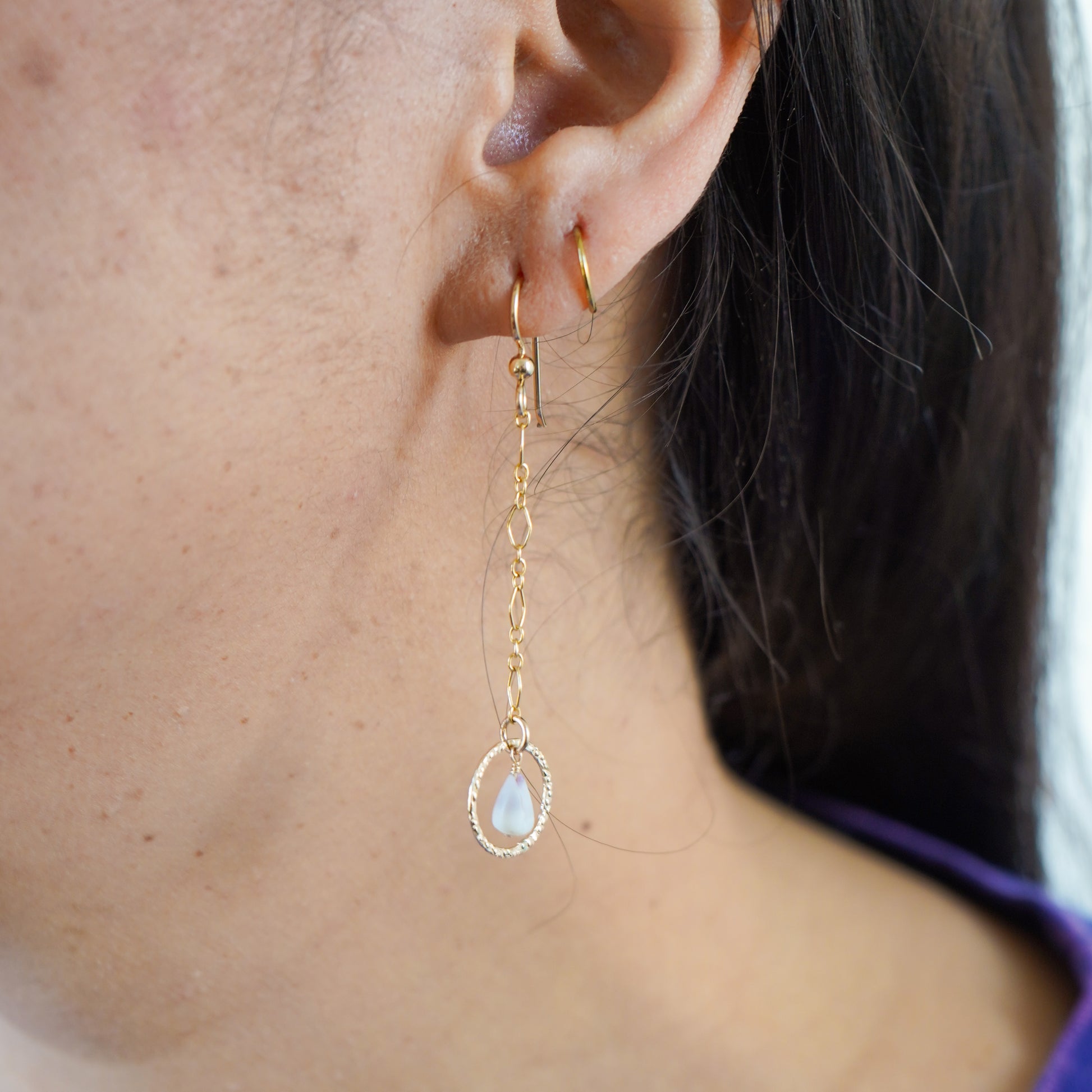 Close-up of a person wearing gold chain dangle earrings with a tiny cone shell.