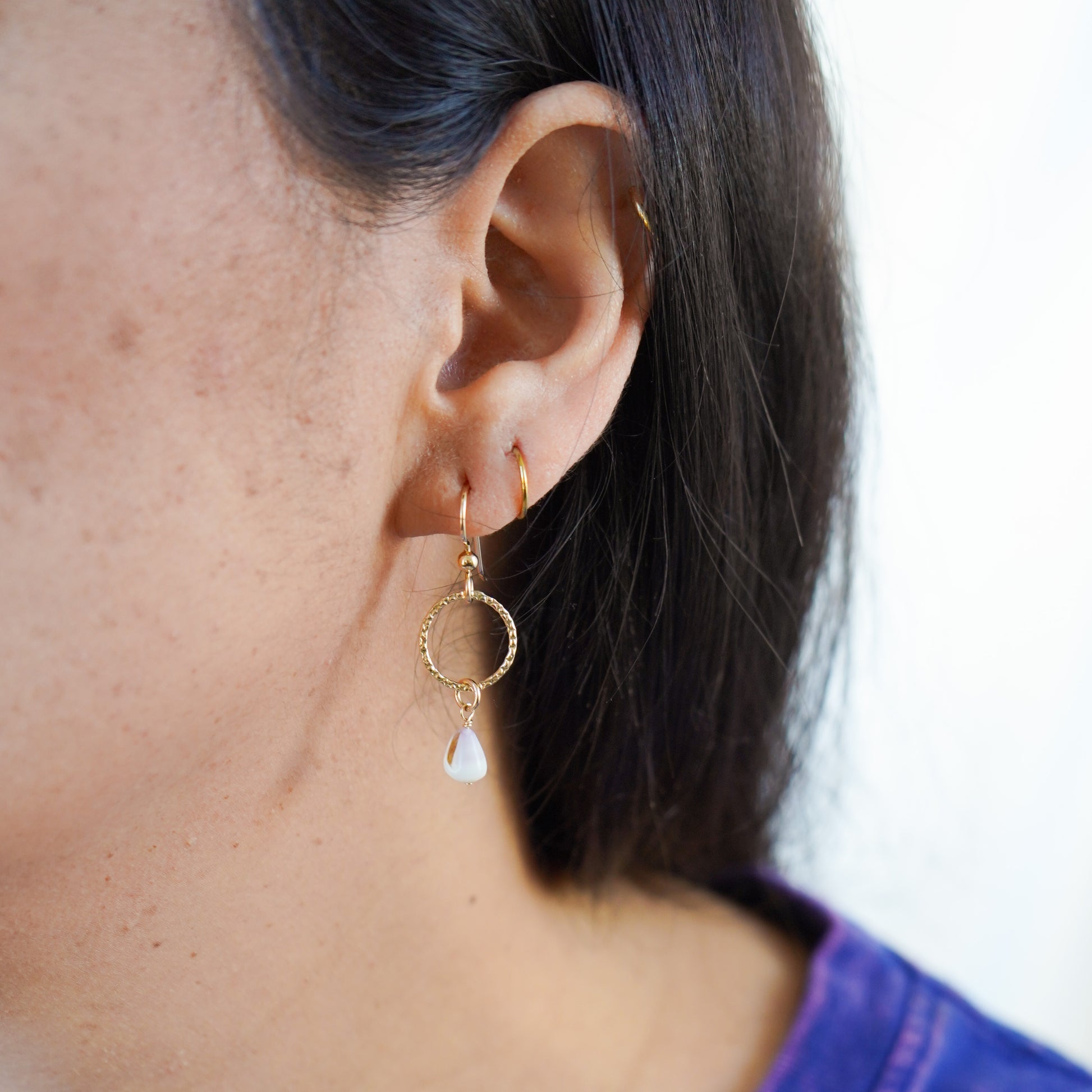 Close-up of an ear wearing gold circle dangle earrings with a tiny white cone shell.