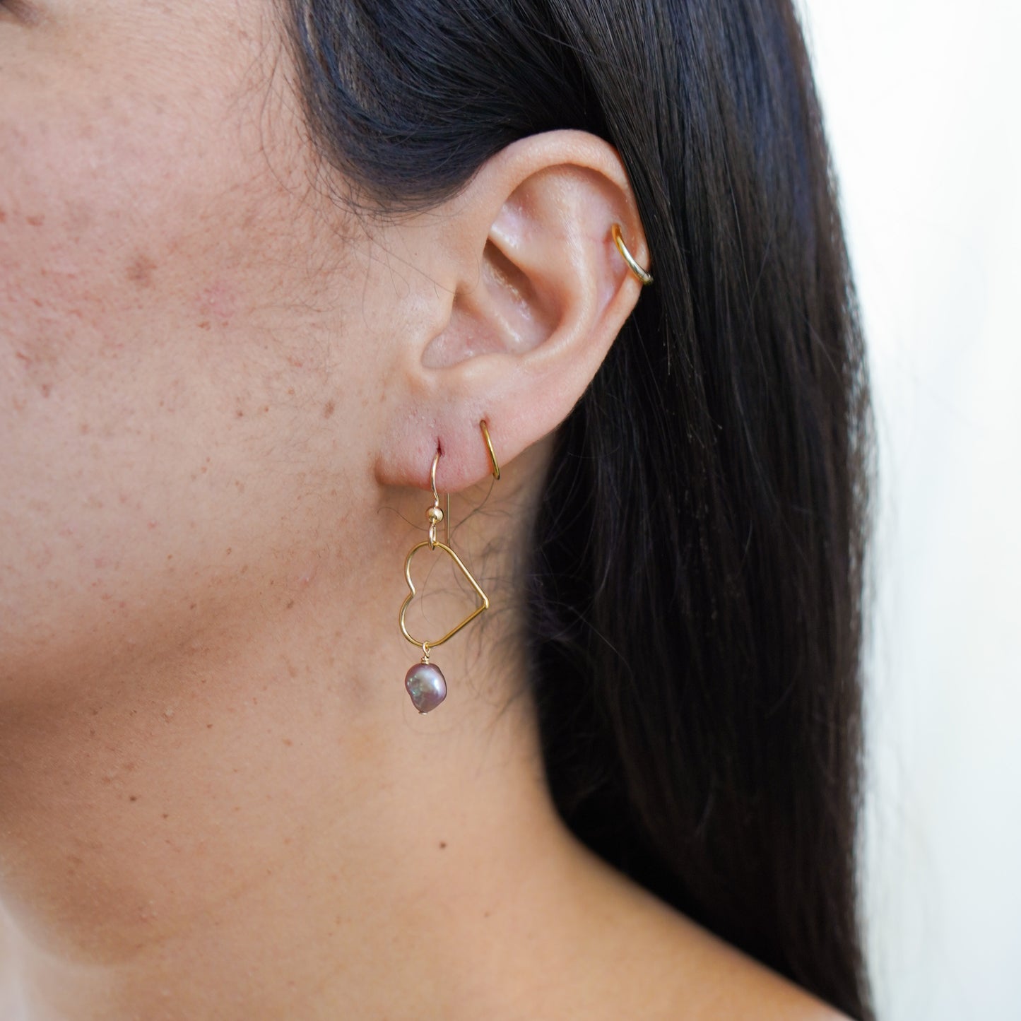 Close-up of an ear wearing gold heart-shaped earrings with pink keshi pearl