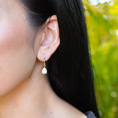 Close-up of an ear wearing a gold earring with a white flower bead, against a blurred green background.