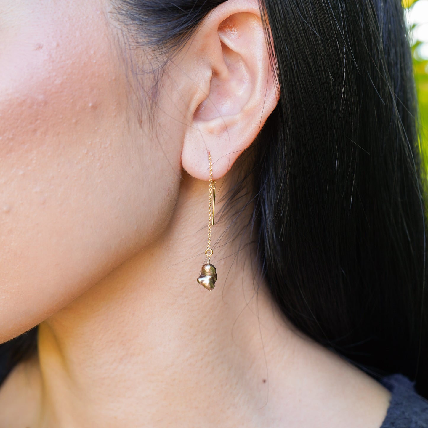 Gold chain dangle earring with brown keshi pearl worn by a person with a blurred background