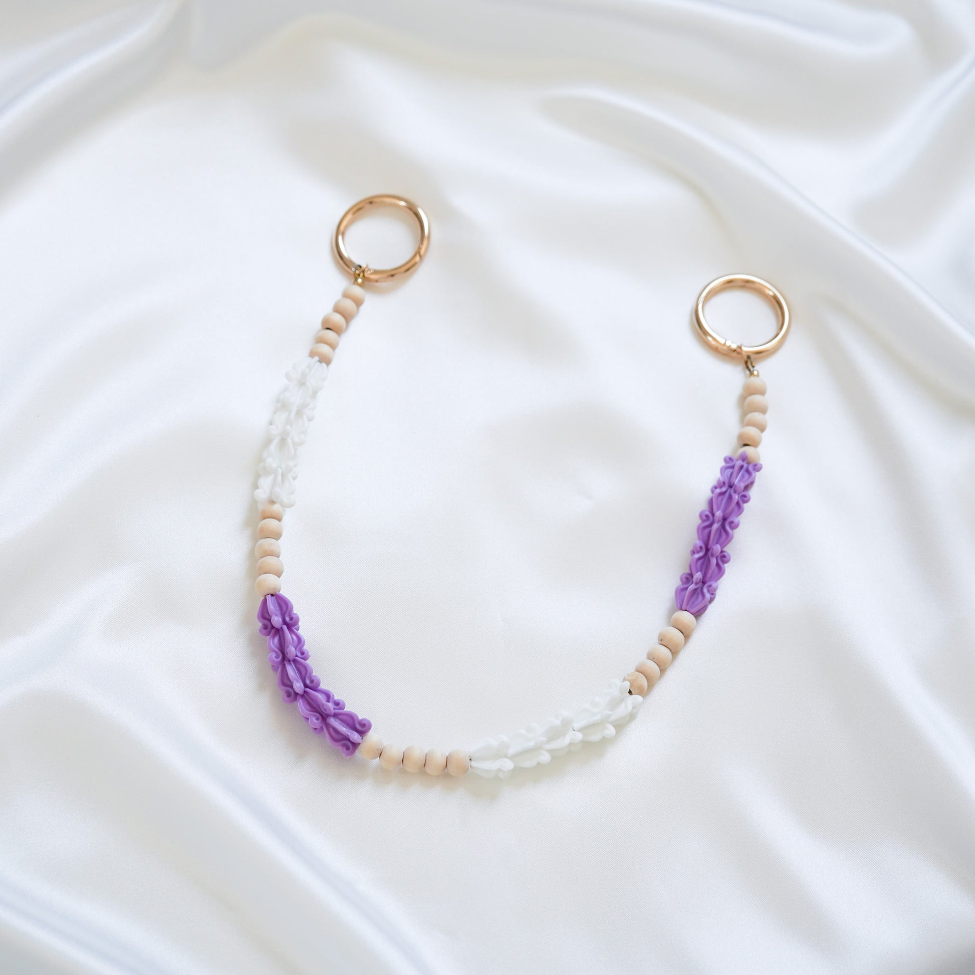 Delicate bag lei featuring purple and white crown flower beads with gold rings on either end on a white fabric background
