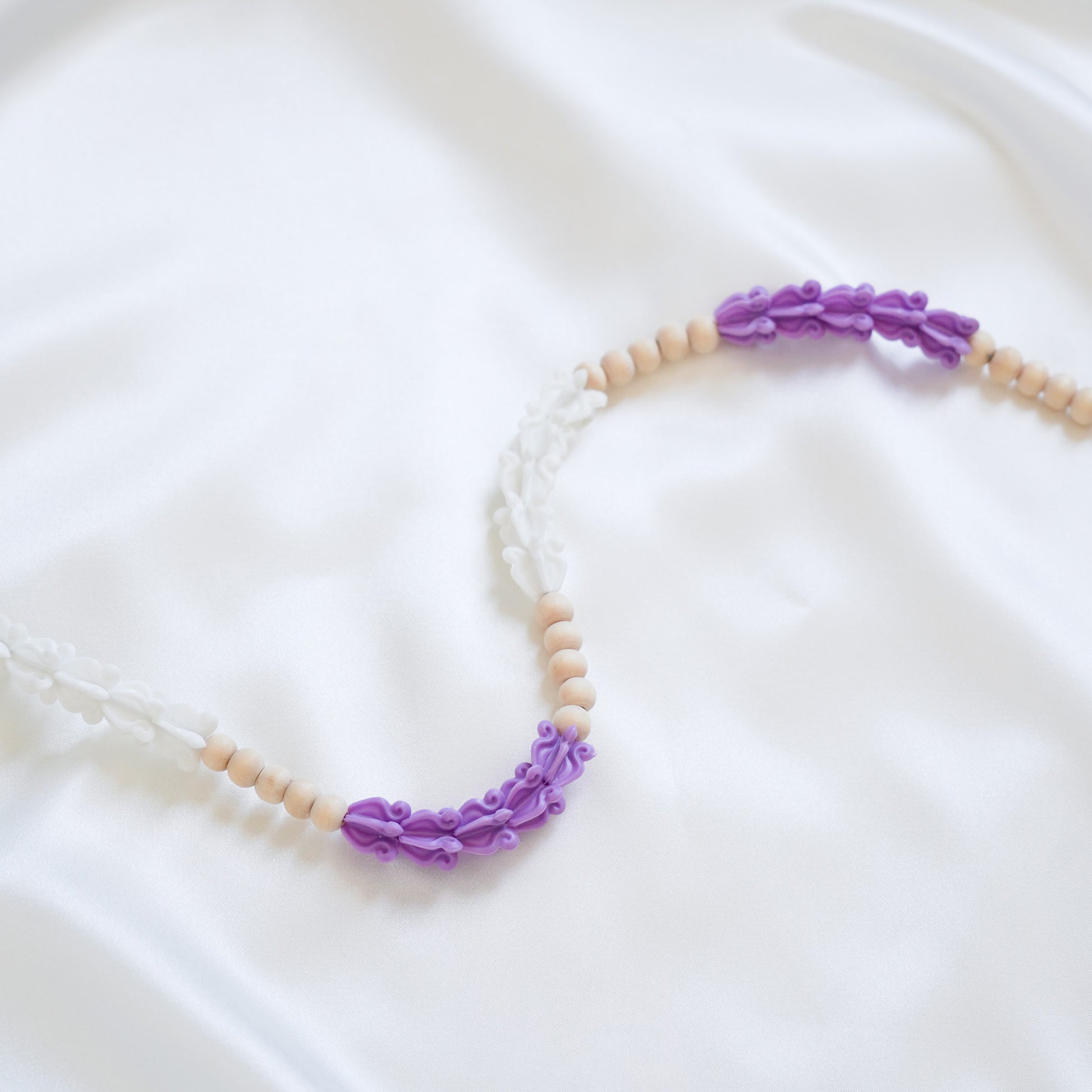 Close up of delicate bag lei featuring purple and white crown flower beads with gold rings on either end on a white fabric background