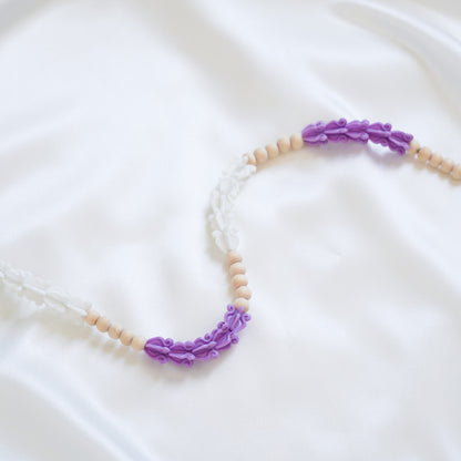 Close up of delicate bag lei featuring purple and white crown flower beads with gold rings on either end on a white fabric background