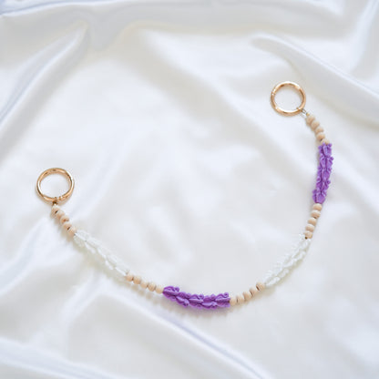 Delicate bag lei featuring purple and white crown flower beads with gold rings on either end on a white fabric background