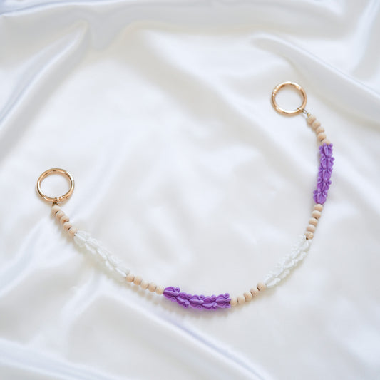 Delicate bag lei featuring purple and white crown flower beads with gold rings on either end on a white fabric background