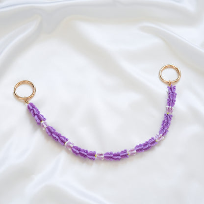 Delicate bag lei featuring purple crown flower beads with gold rings on either end on a white fabric background