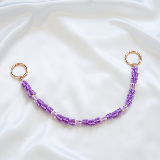 Delicate bag lei featuring purple crown flower beads with gold rings on either end on a white fabric background