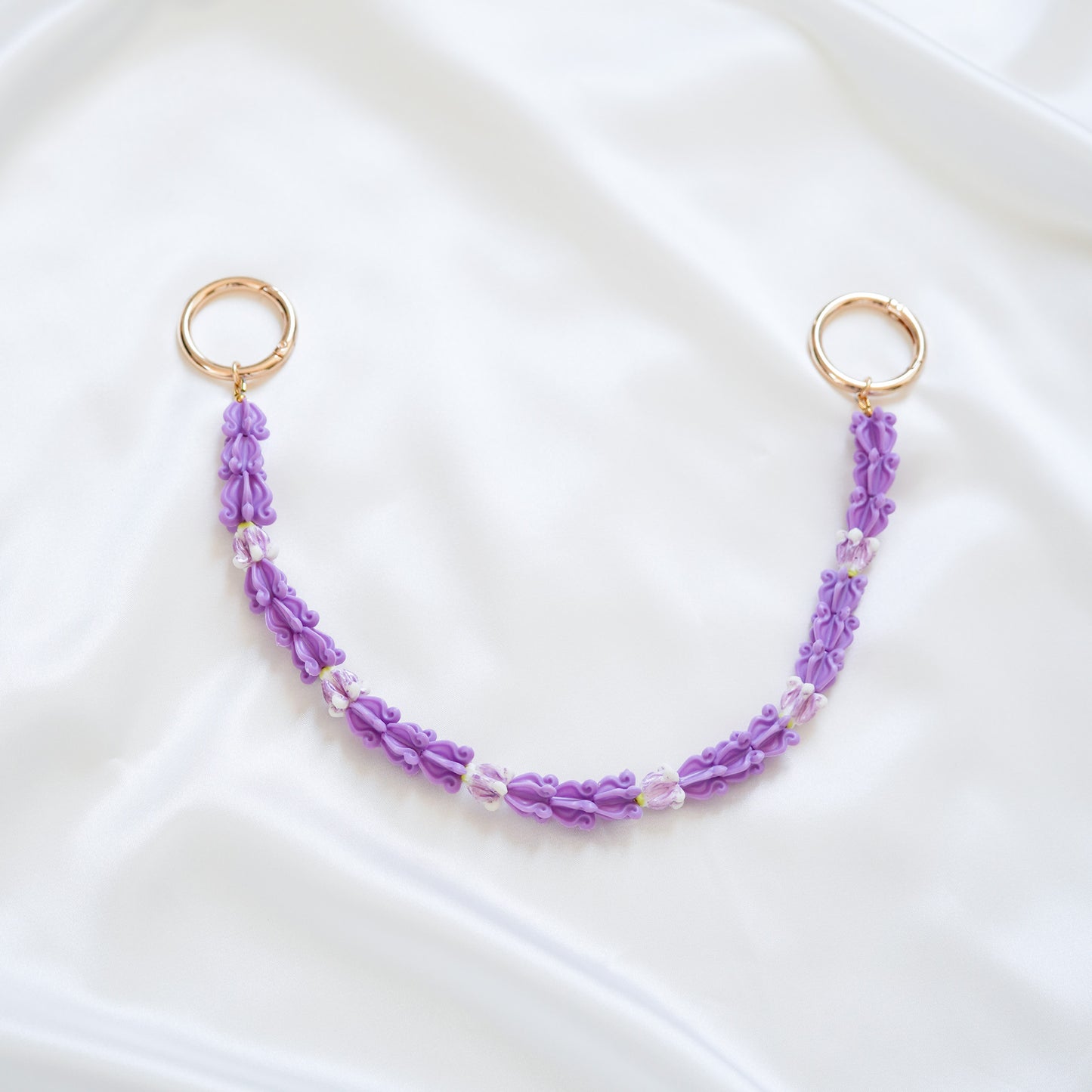 Delicate bag lei featuring purple crown flower beads with gold rings on either end on a white fabric background