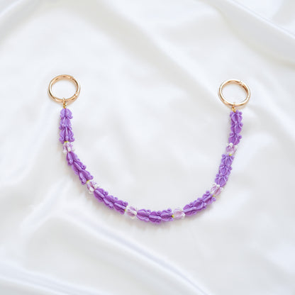 Delicate bag lei featuring purple crown flower beads with gold rings on either end on a white fabric background