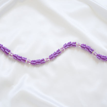 Close up of delicate bag lei featuring purple crown flower beads with gold rings on either end on a white fabric background