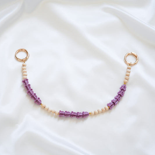 Delicate bag lei featuring alternating purple crown flower beads and tan beads with gold rings on either end on a white fabric background