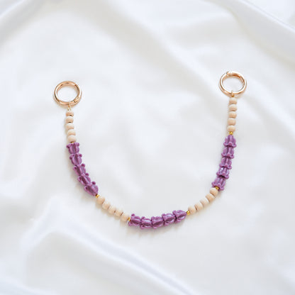 Delicate bag lei featuring alternating purple crown flower beads and tan beads with gold rings on either end on a white fabric background