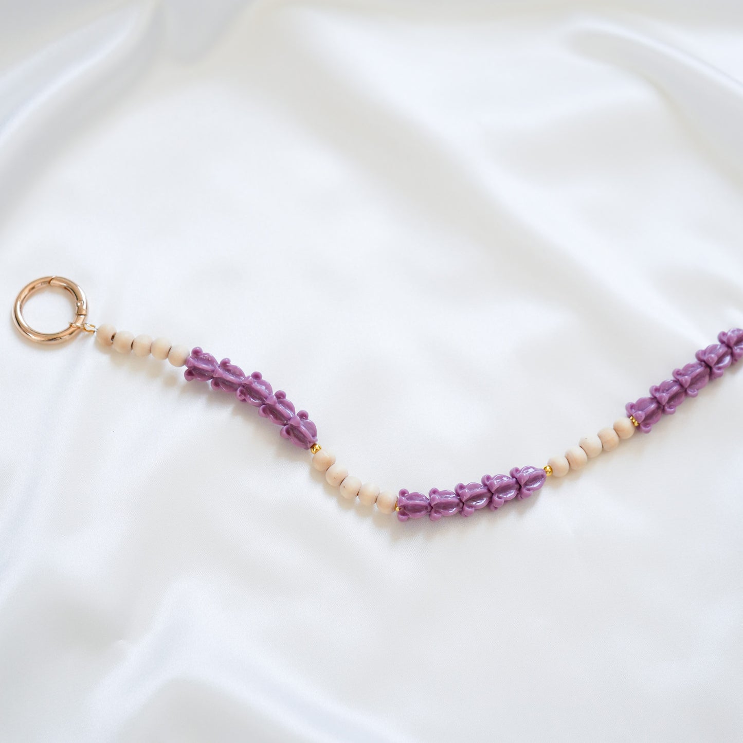 Close up of delicate bag lei featuring alternating purple crown flower beads and tan beads with gold rings on either end on a white fabric background