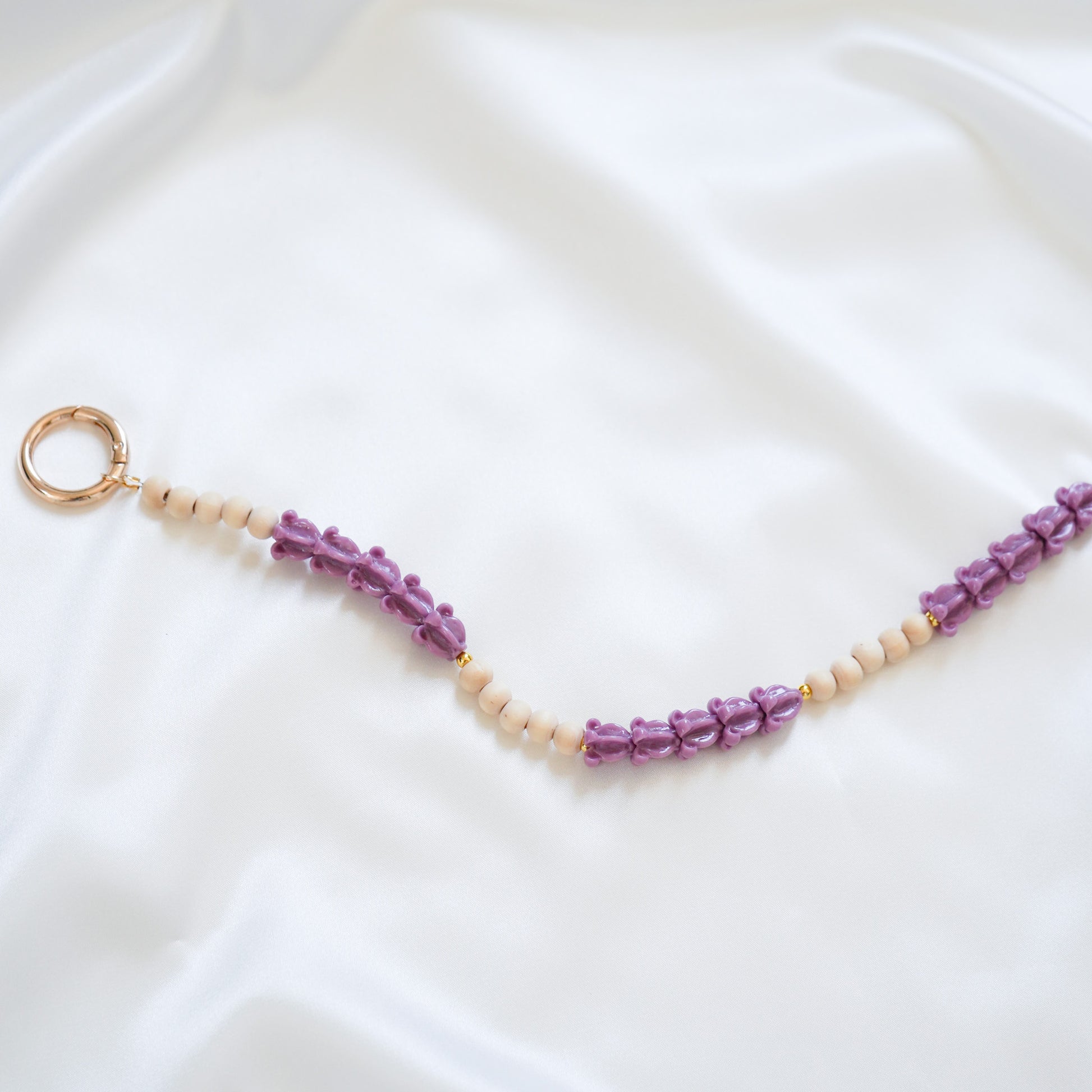 Close up of delicate bag lei featuring alternating purple crown flower beads and tan beads with gold rings on either end on a white fabric background