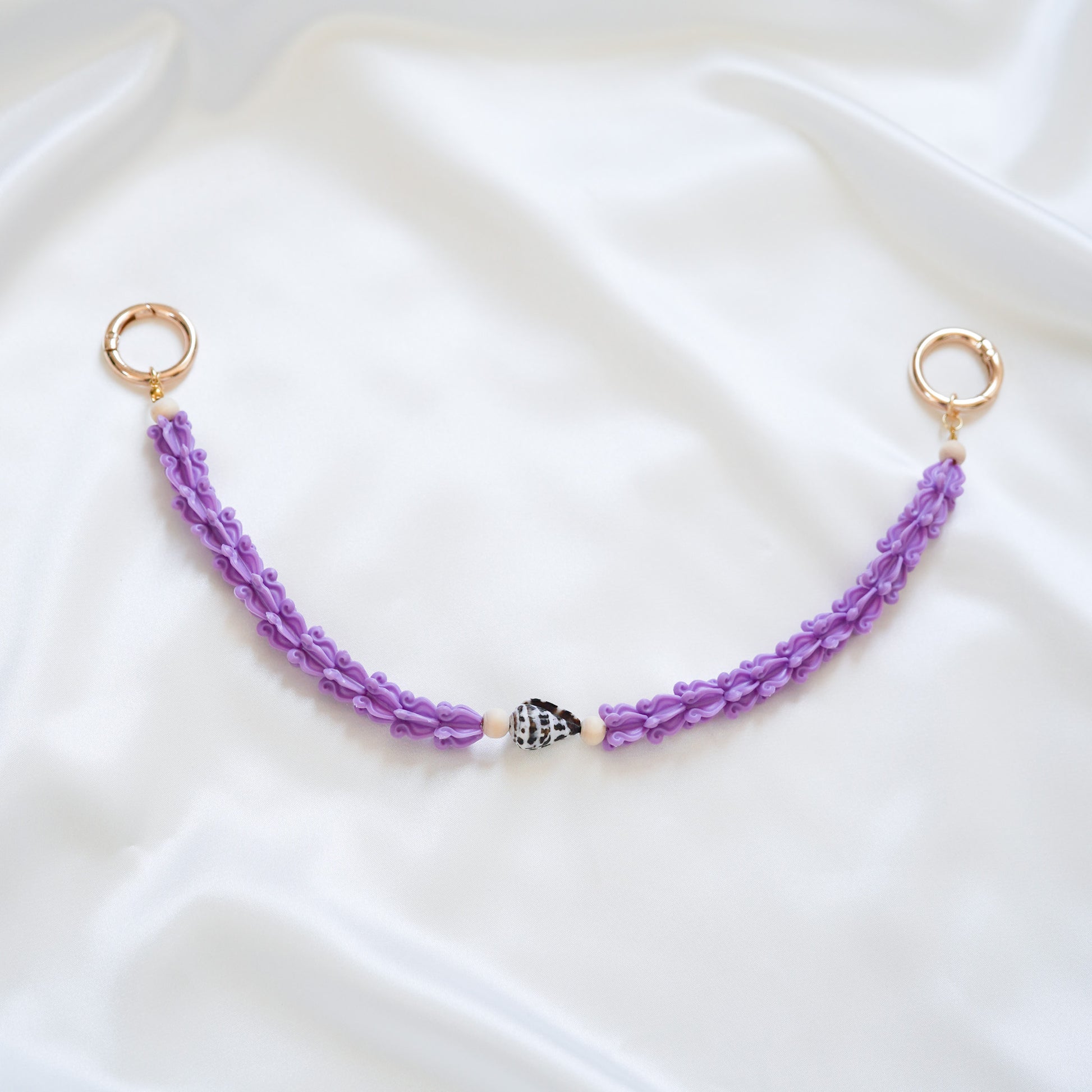 Delicate bag lei featuring purple crown flower beads with brown and white hebrew shell in middle with gold rings on either end on a white fabric background