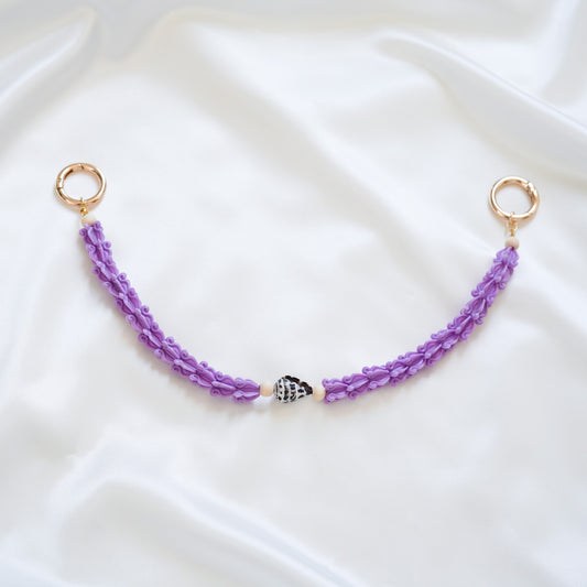 Delicate bag lei featuring purple crown flower beads with brown and white hebrew shell in middle with gold rings on either end on a white fabric background