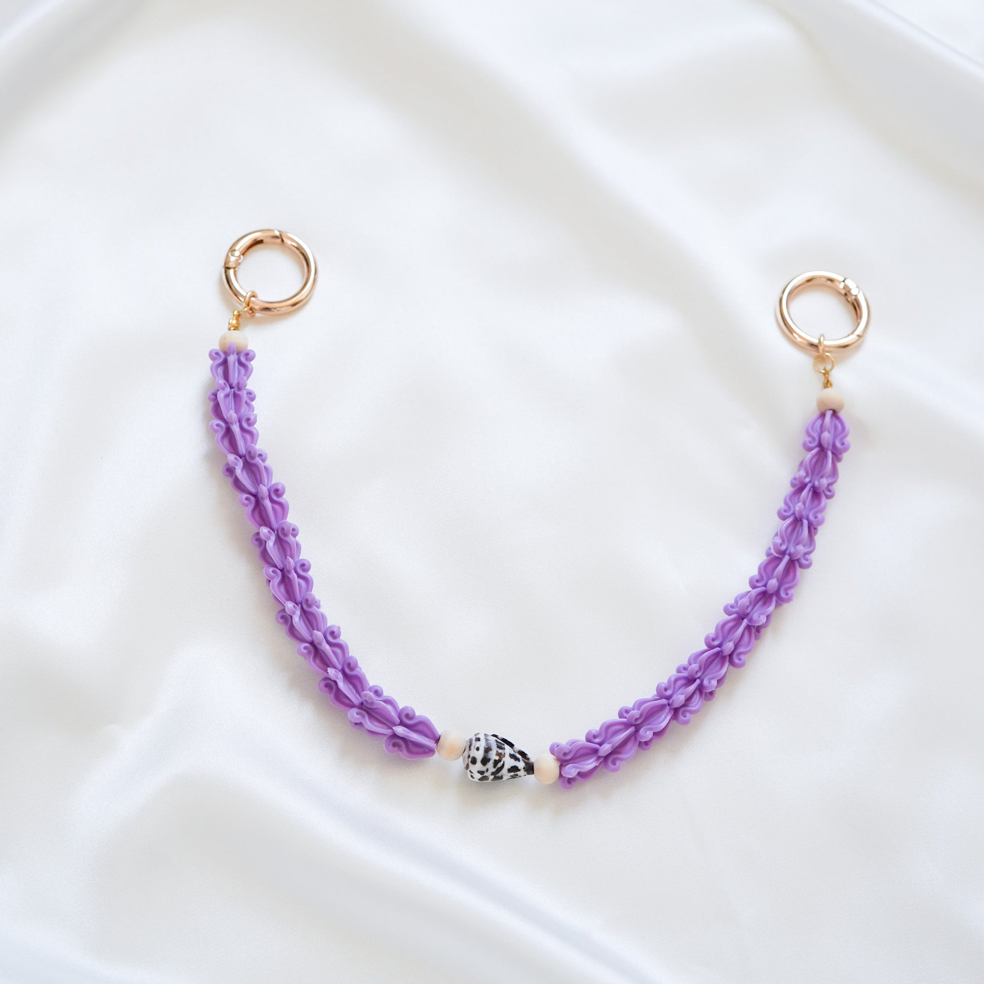 Delicate bag lei featuring purple crown flower beads with brown and white hebrew shell in middle with gold rings on either end on a white fabric background