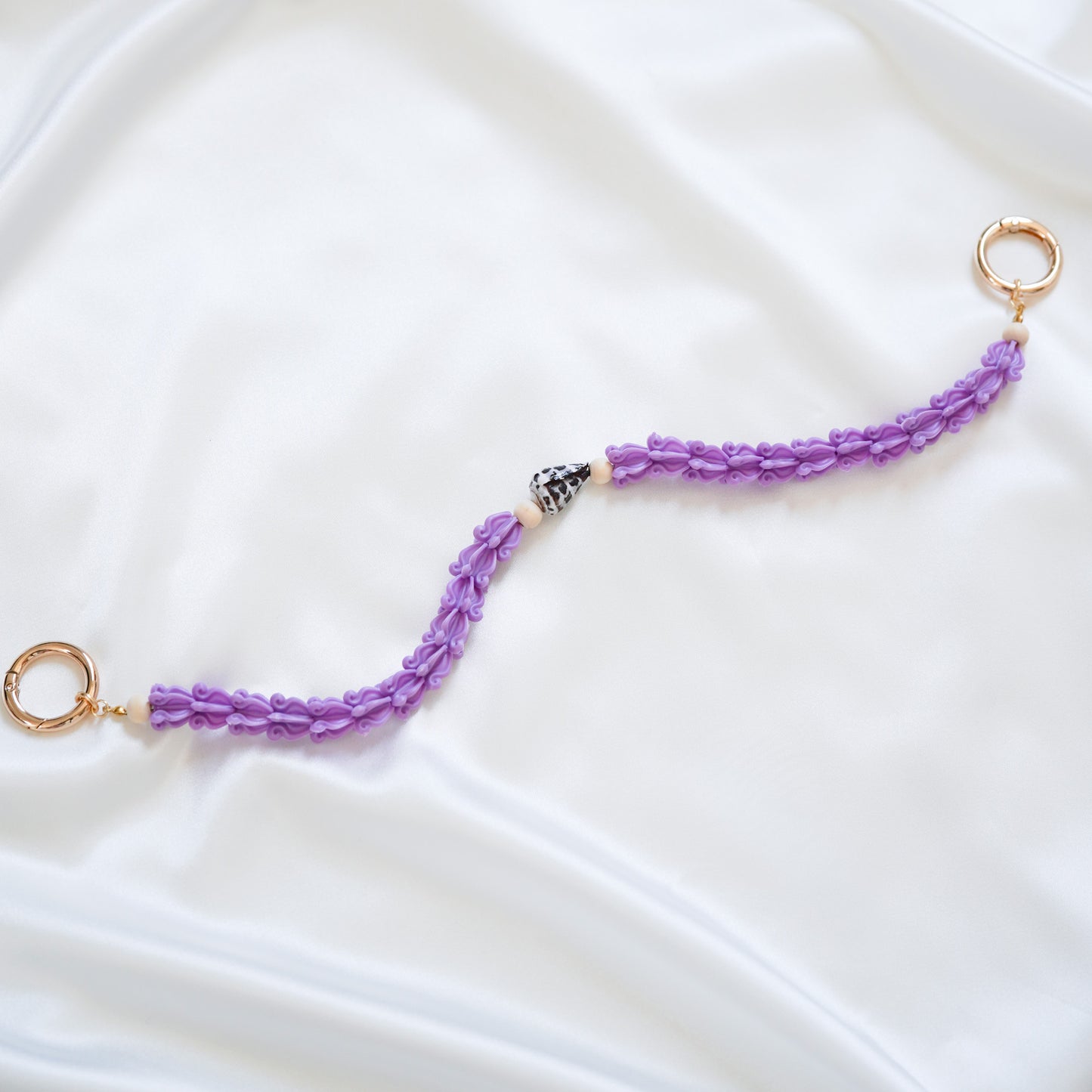 Delicate bag lei featuring purple crown flower beads with brown and white hebrew shell in middle with gold rings on either end on a white fabric background