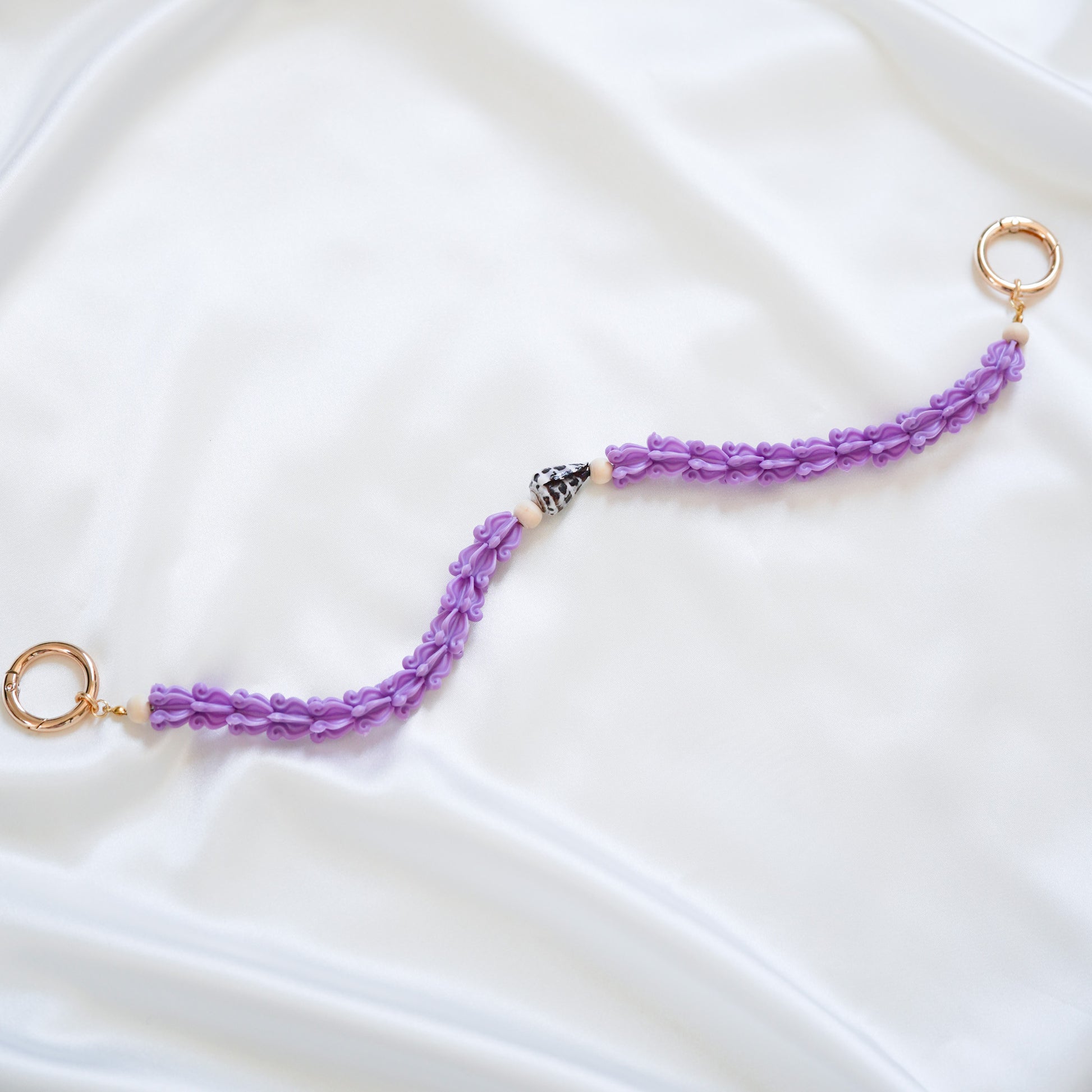 Delicate bag lei featuring purple crown flower beads with brown and white hebrew shell in middle with gold rings on either end on a white fabric background