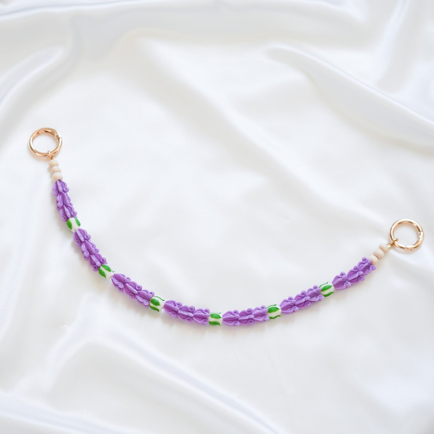 Delicate bag lei featuring purple crown flower beads with white rose beads with gold rings on either end on a white fabric background
