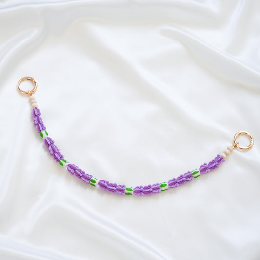 Delicate bag lei featuring purple crown flower beads with white rose beads with gold rings on either end on a white fabric background