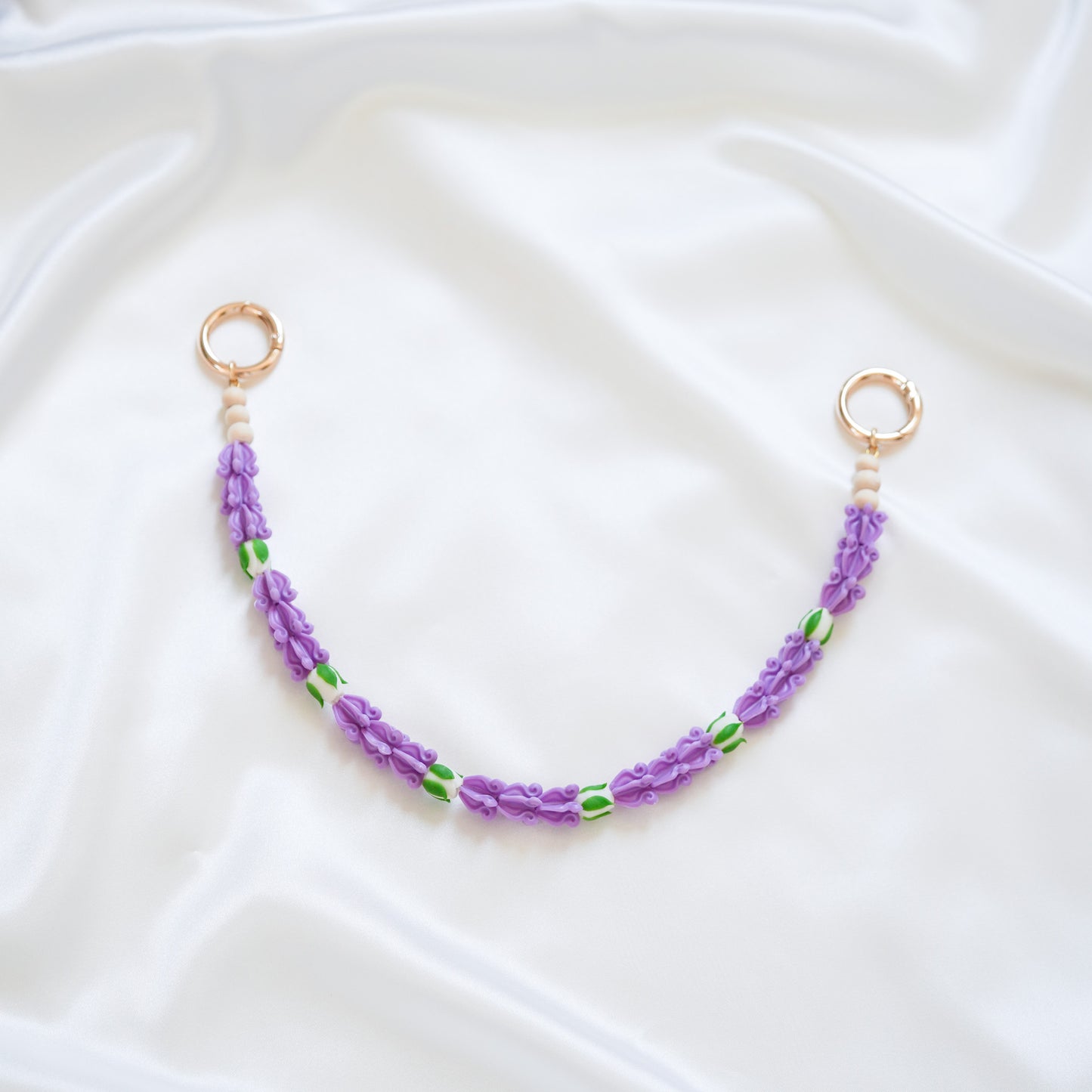 Delicate bag lei featuring purple crown flower beads with white rose beads with gold rings on either end on a white fabric background