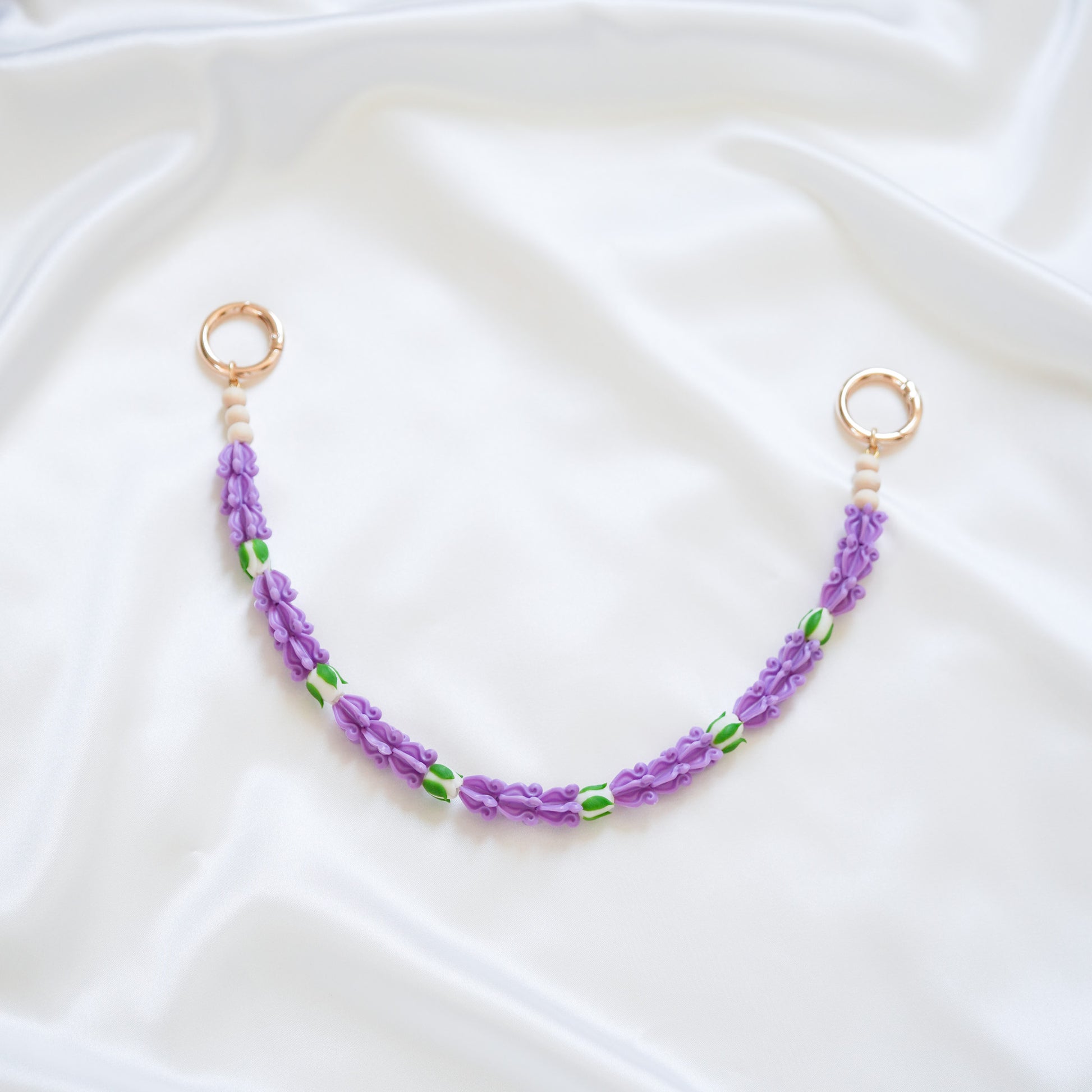 Delicate bag lei featuring purple crown flower beads with white rose beads with gold rings on either end on a white fabric background