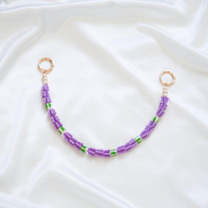 Delicate bag lei featuring purple crown flower beads with white rose beads with gold rings on either end on a white fabric background