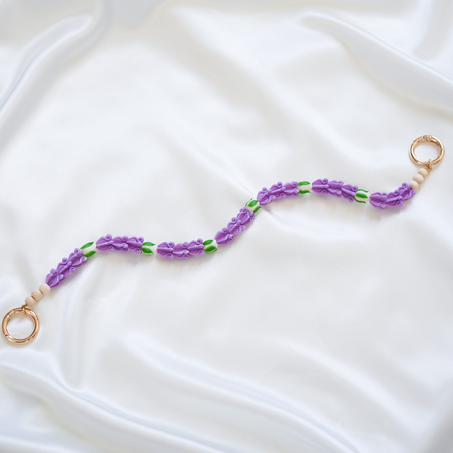 Delicate bag lei featuring purple crown flower beads with white rose beads with gold rings on either end on a white fabric background