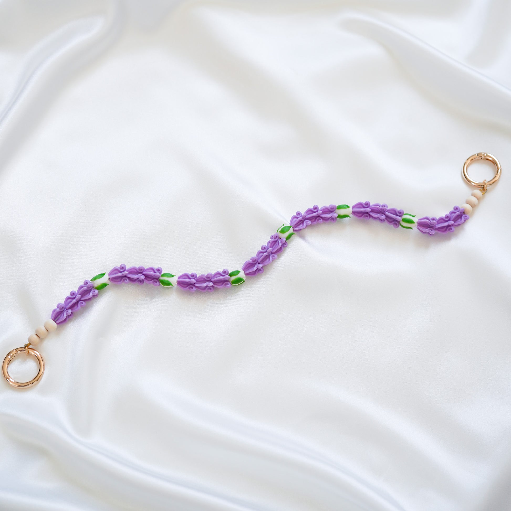 Delicate bag lei featuring purple crown flower beads with white rose beads with gold rings on either end on a white fabric background