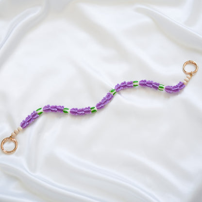 Delicate bag lei featuring purple crown flower beads with white rose beads with gold rings on either end on a white fabric background