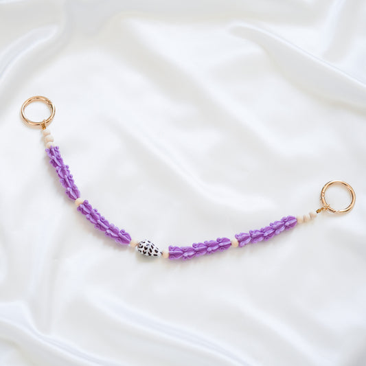 Delicate bag lei featuring purple crown flower beads with white and brown hebrew shell and tan bead spacer beads with gold rings on either end on a white fabric background