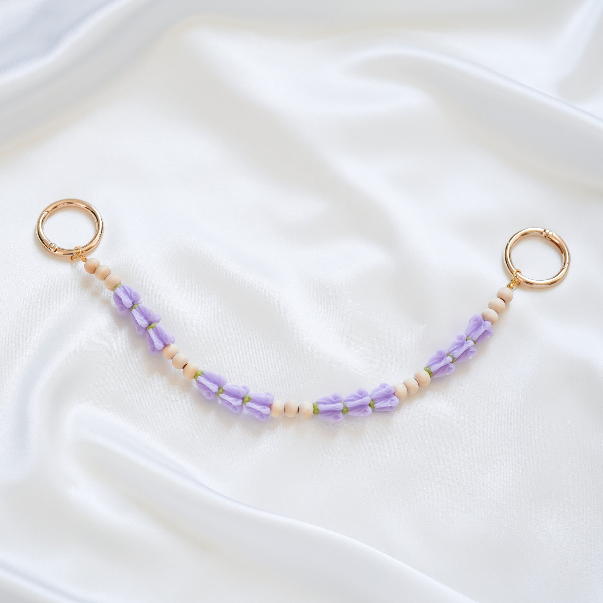 Delicate bag lei featuring puprle crown flower and tan beads in sets of three with gold rings on either end on a white fabric background