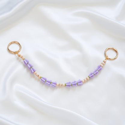 Delicate bag lei featuring puprle crown flower and tan beads in sets of three with gold rings on either end on a white fabric background